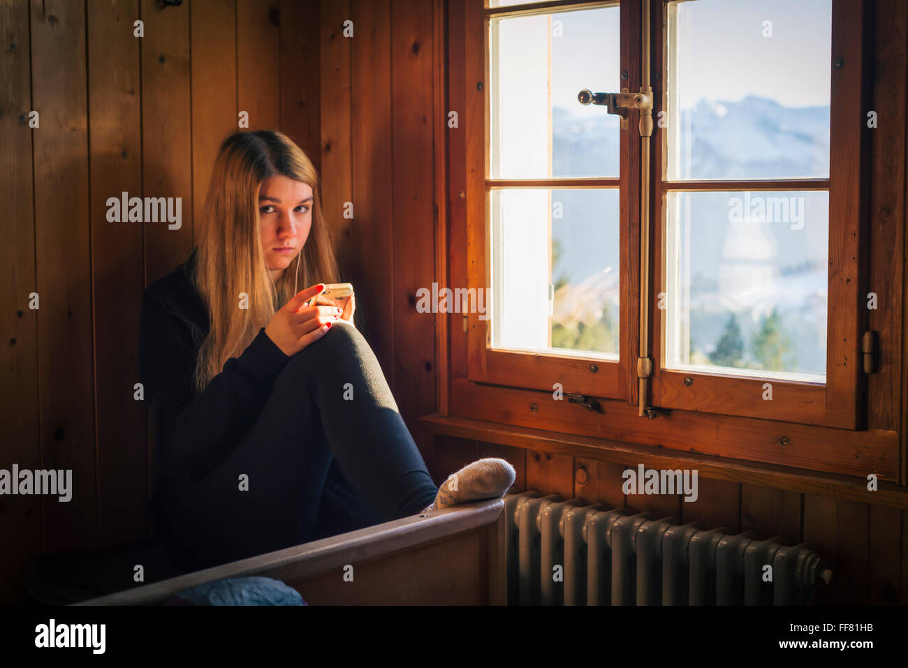 Junge Frau mit Smartphone in Kamera sitzt im Inneren Berghütte mit Holzverkleidung Wand hinter dem Fenster Stockfoto