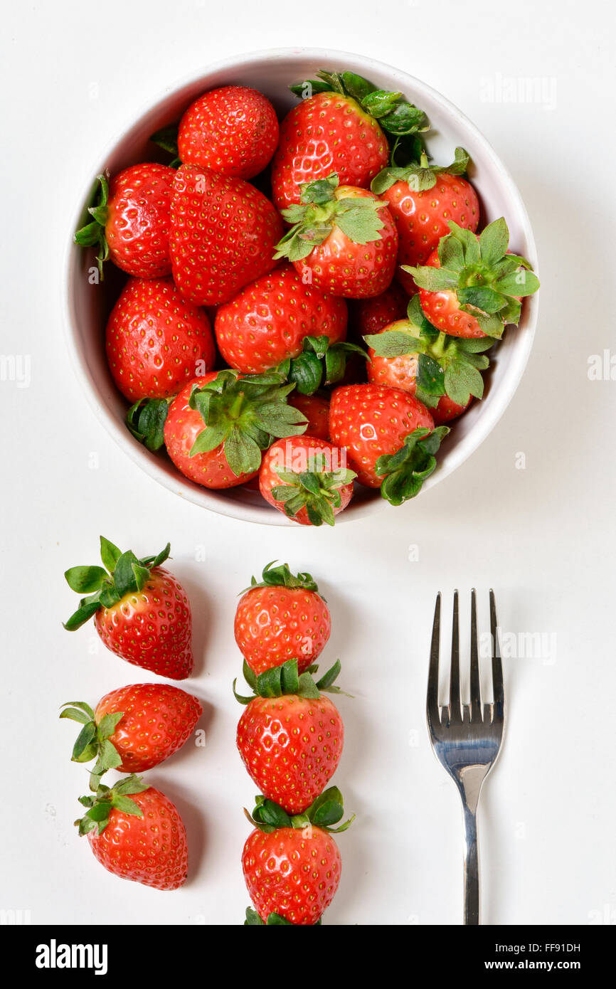 High-Angle Shot einer weißen Keramik Schüssel voll appetitlich Erdbeeren, eine Gabel und einige andere Erdbeeren auf einer weißen Fläche Stockfoto