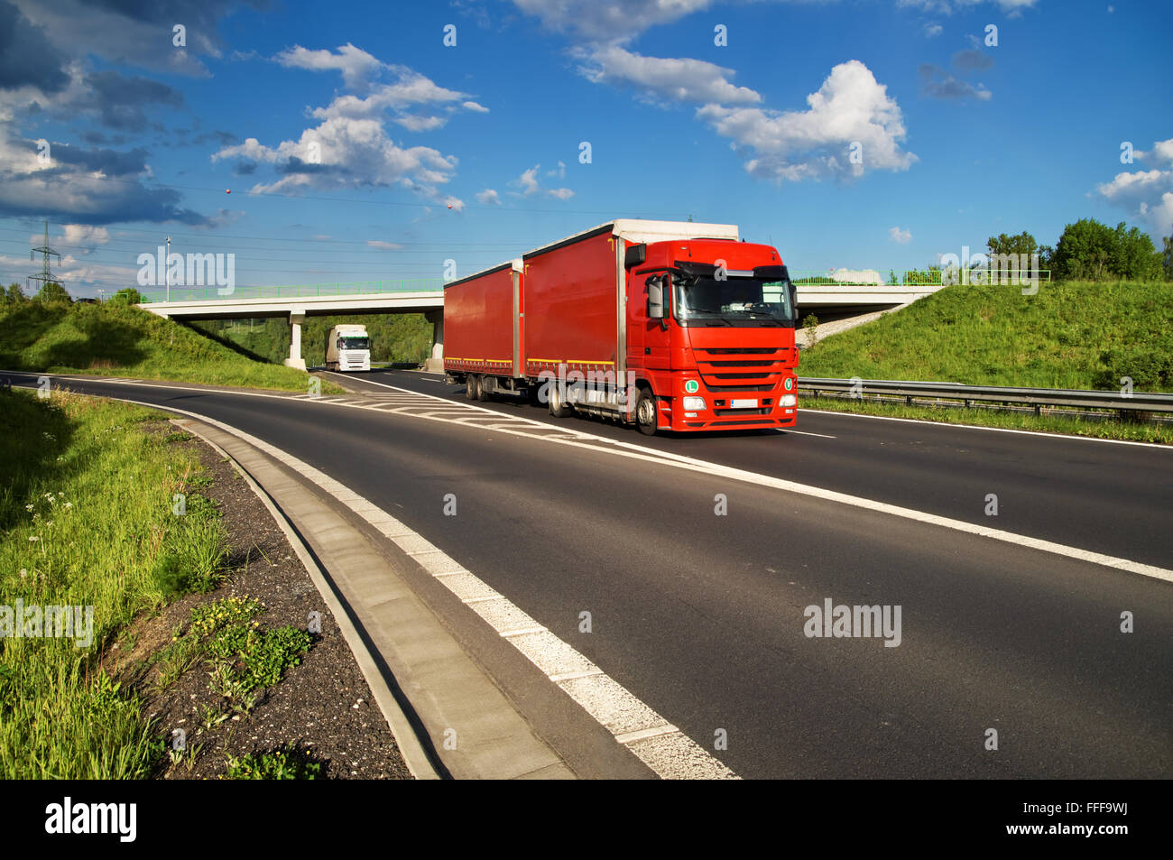 Rote LKW-fahren auf dem Highway durch die Landschaft, im Hintergrund die Brücke unter verläuft einen weißen LKW Stockfoto