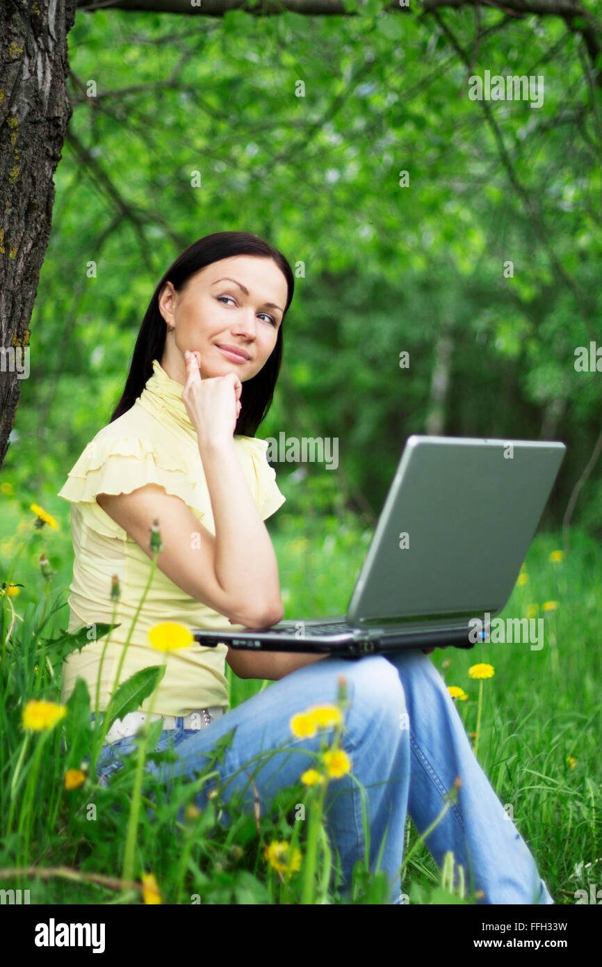 Junge Frau mit Laptop auf den Knien auf dem Rasen sitzen und denken über die Zukunft oder Vergangenheit. Stockfoto