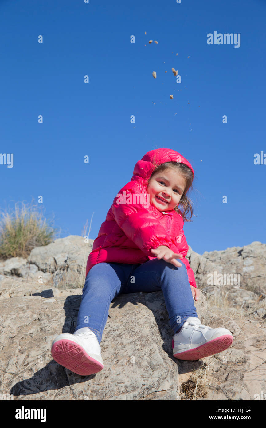 Schöne kleine Mädchen werfen Steinchen, Kamera auf Felsen sitzend Stockfoto