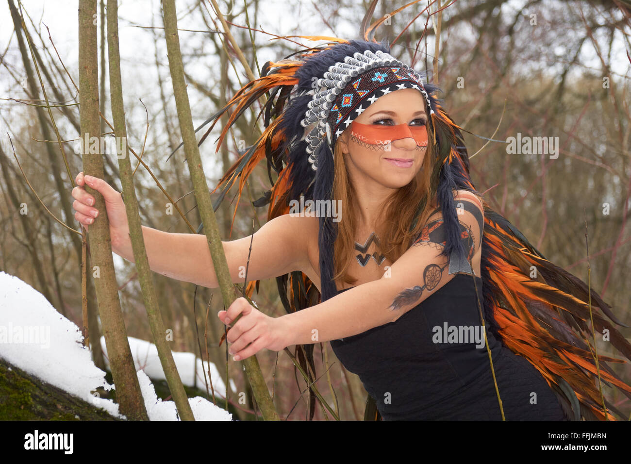 Mädchen in Indianer Kopfschmuck auf dem Baum im Winterwald Stockfoto