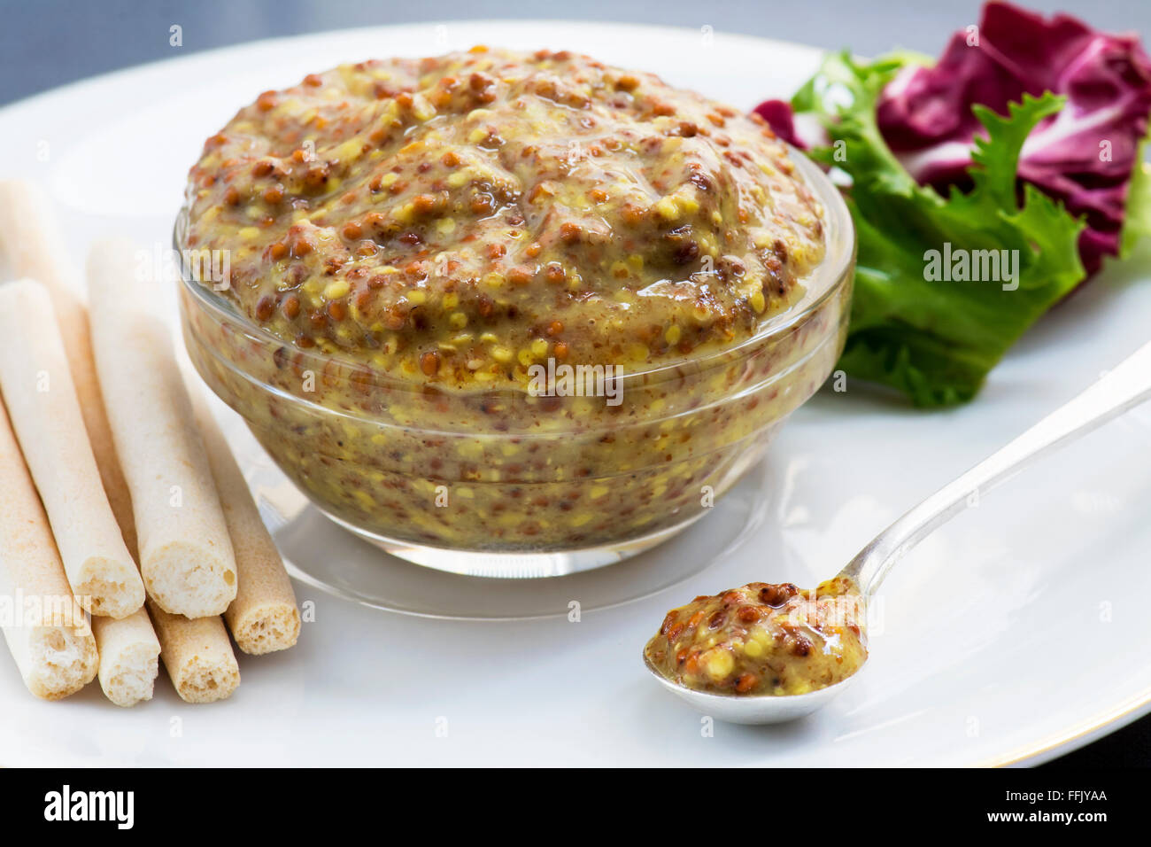 Dijon-Senf mit Brot-Sticks und Salat. Fokus auf vorderen Senf in einem kleinen silbernen Löffel auf einem weißen Porzellanteller Stockfoto