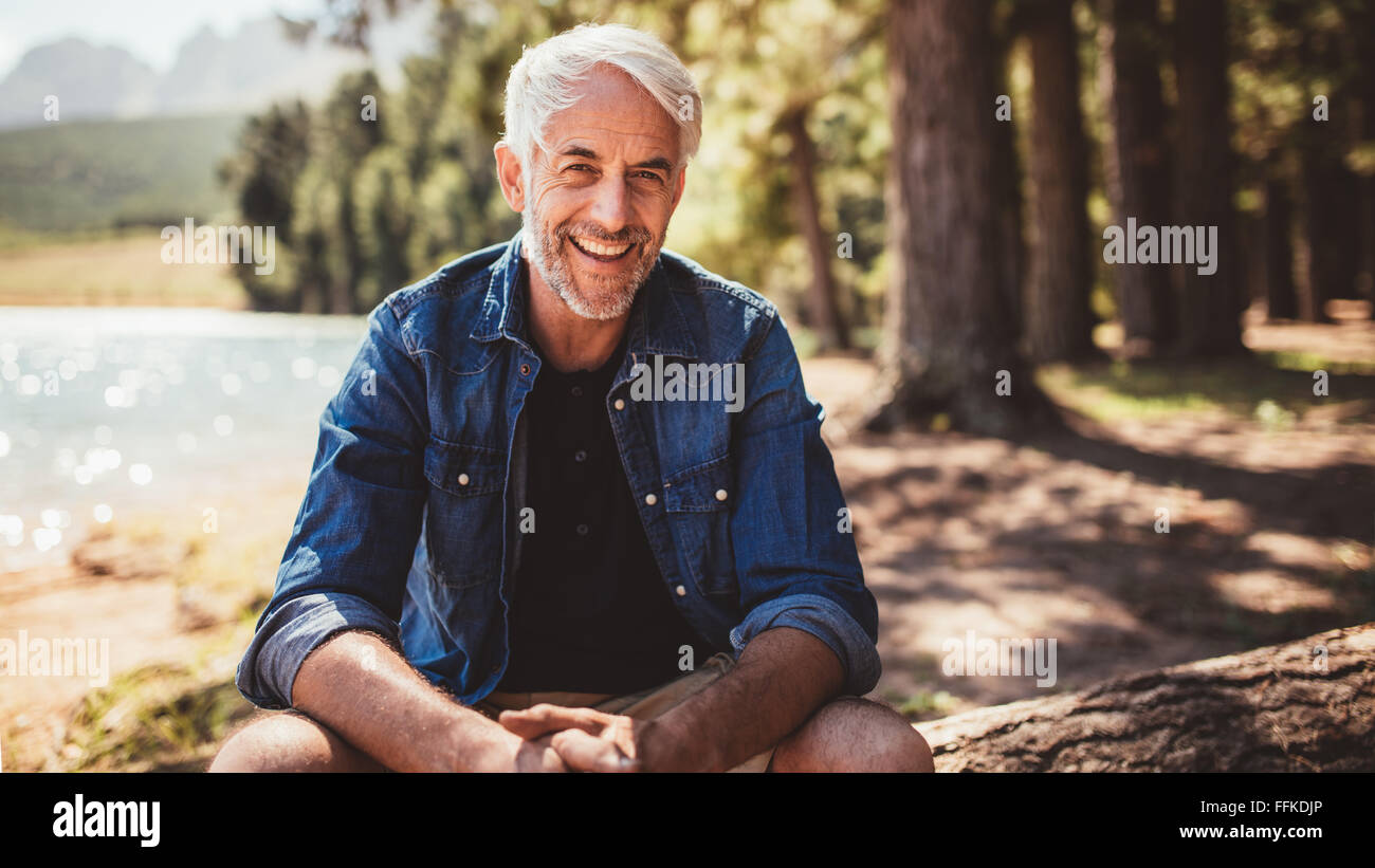 Porträt von glücklich reifer Mann sitzt in der Nähe von einem See Blick in die Kamera und lächelnd. Senior kaukasischen Mann sitzt auf einem Baumstamm von der lak Stockfoto