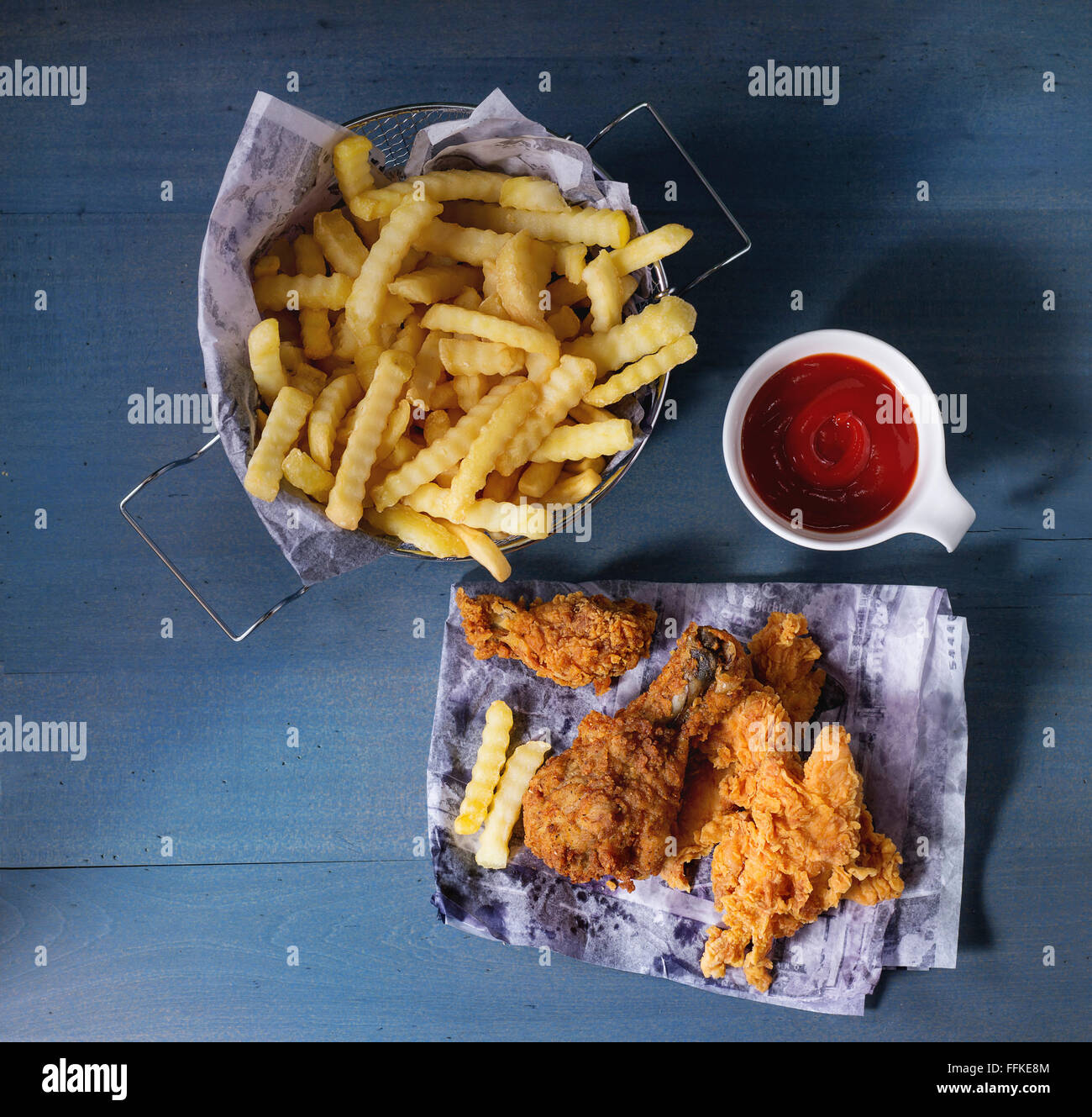 Chicken Pommes Streifen und Beine auf dem Papier mit Korb mit Pommes Frites und Schale mit Ketchup-Sauce über blaue Holztisch. Ansicht von oben. Stockfoto