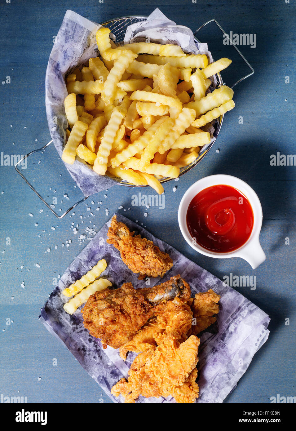 Chicken Pommes Streifen und Beine auf dem Papier mit Korb mit Pommes Frites und Schale mit Ketchup-Sauce über blaue Holztisch. Ansicht von oben Stockfoto