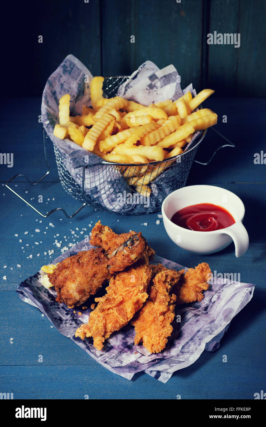 Chicken Pommes Streifen und Beine auf dem Papier mit Korb mit Pommes Frites und Schale mit Ketchup-Sauce über blaue Holztisch. Türkis Stockfoto