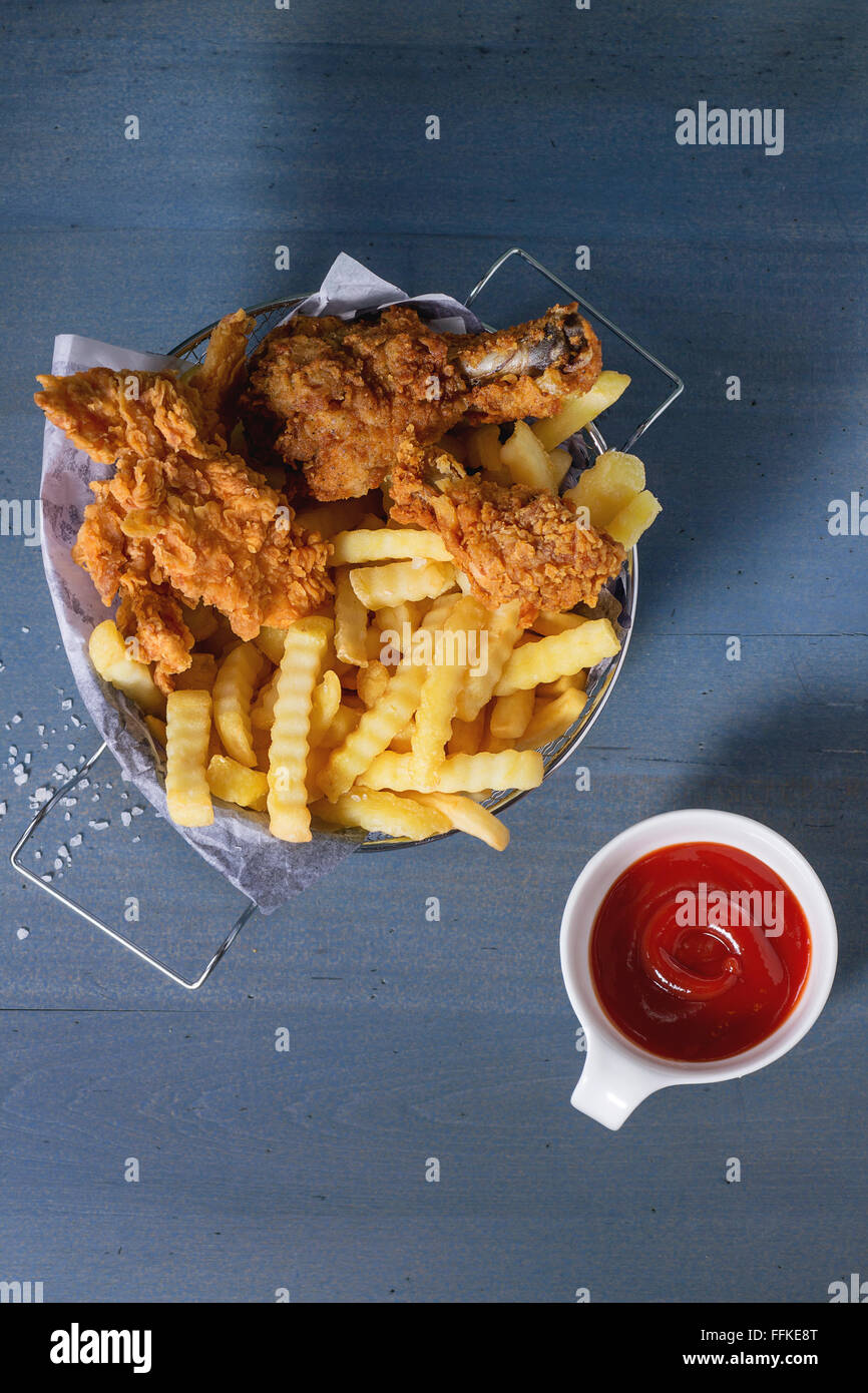 Chicken Pommes Streifen und Beine mit Pommes frites im Metallkorb und Schüssel mit Ketchup-Sauce über blaue Holztisch mit Meersalz. Stockfoto