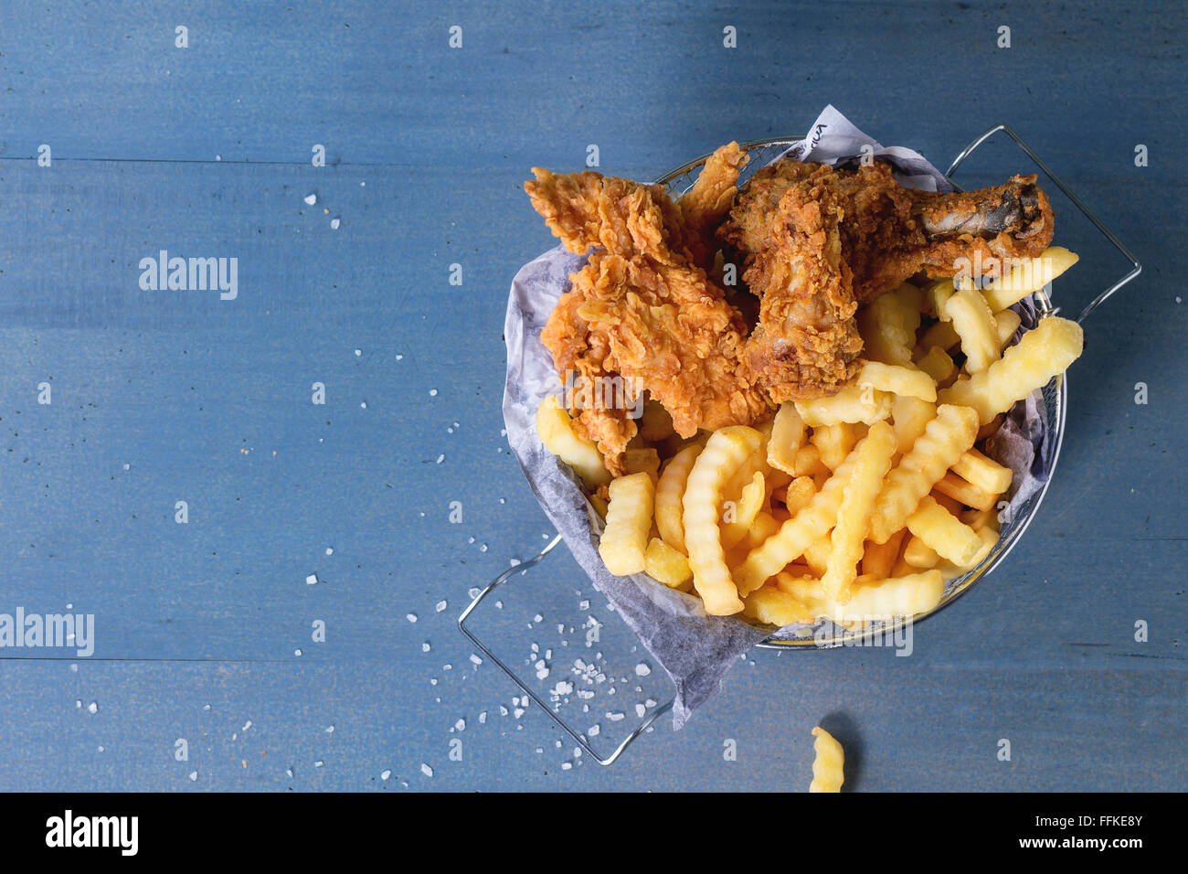 Pommes frites Hähnchenstreifen und Beine mit Pommes frites im Metallkorb über blaue Holztisch mit Meersalz. Ansicht von oben Stockfoto