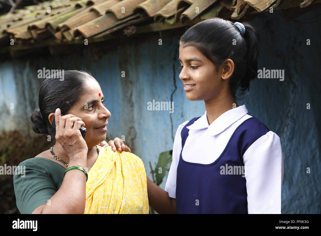 Mutter am Handy und Schule gehen Tochter vor Haus im Dorf, Maharashtra, Indien Model Release # 703Y und 703Z sprechen Stockfoto
