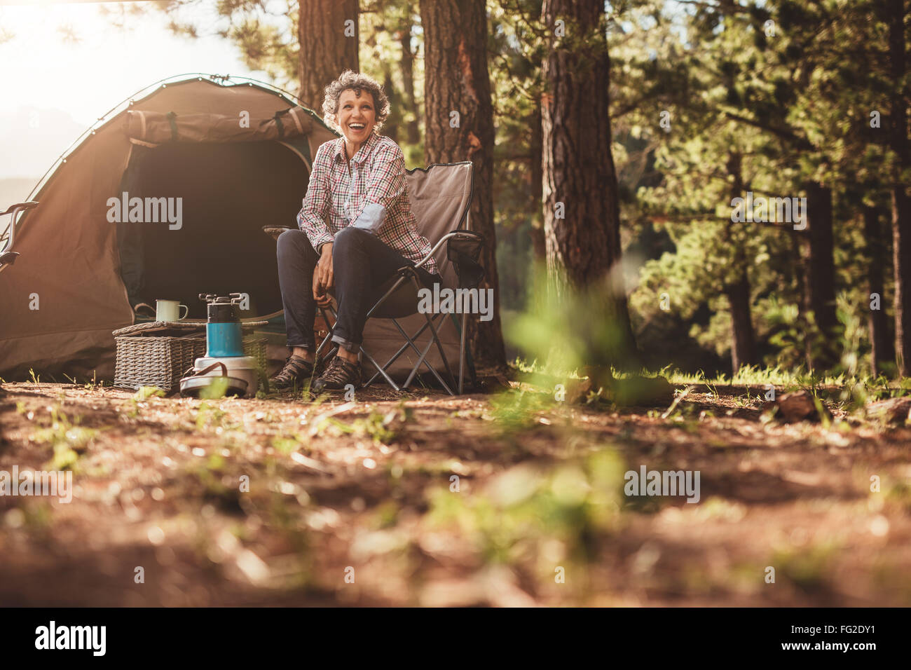 Ältere Frau sitzt entspannt vor einem Zelt. Reife Frau im Wald camping lächelnd. Stockfoto