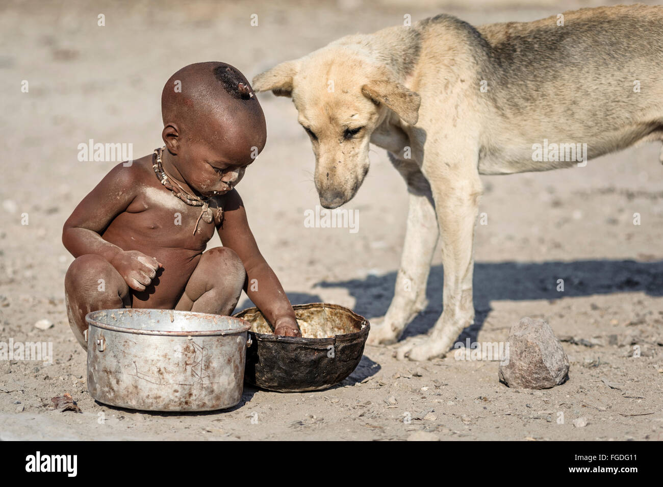 Himba Kind sitzt auf dem Boden sein entlegenes Dorf aus einem rostigen Topf Essen, während ein Hund in seiner Nähe sucht. Stockfoto