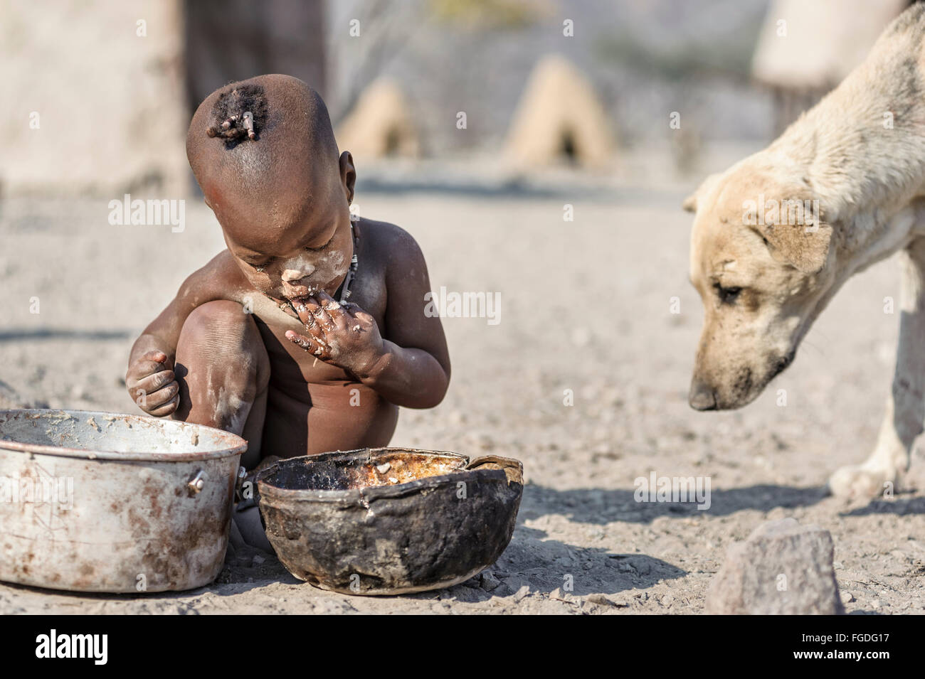 Himba Kind sitzt auf dem Boden sein entlegenes Dorf aus einem rostigen Topf Essen, während ein Hund in seiner Nähe sucht. Stockfoto