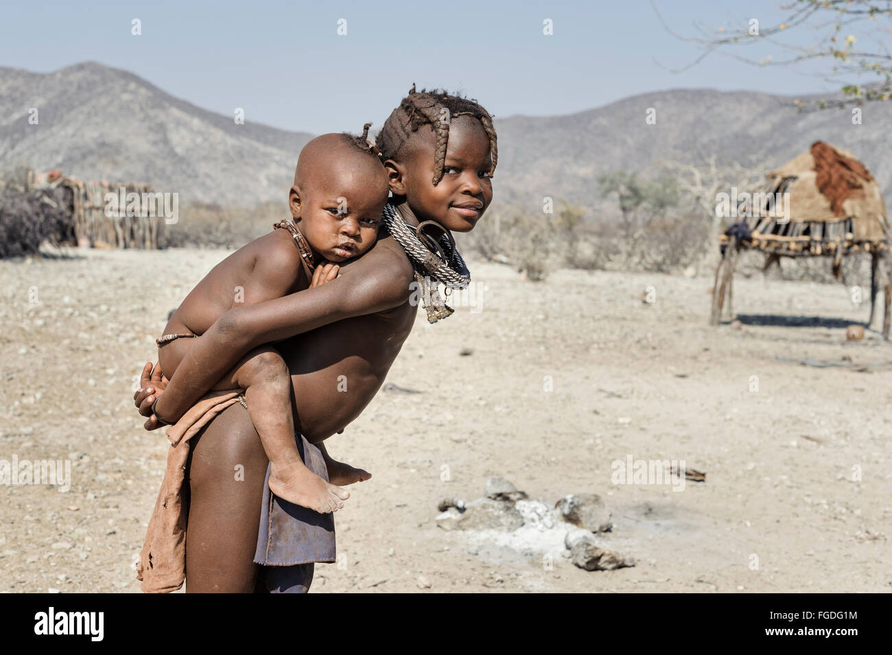 Himba-Kind mit ihrer Schwester auf dem Rücken um ihren abgelegenen Dorf. Stockfoto