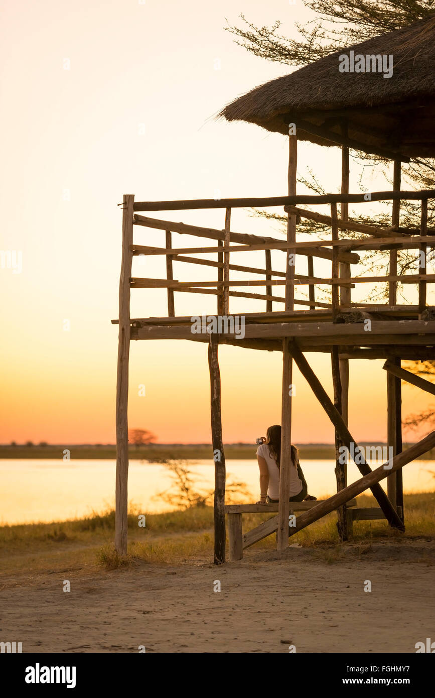 Ein junger weiblicher Tourist sitzt unter einem Strohdach-Hütte und macht Fotos von Sonnenuntergang Weile auf Safari in die Makgadikgadi Salzpfannen, Botsw Stockfoto