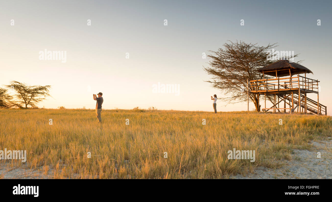 Ein junges Paar lange Gras stehen und Fotos von Sonnenuntergang Weile auf Safari in die Makgadikgadi Salzpfannen, Botswana, Afrika Stockfoto