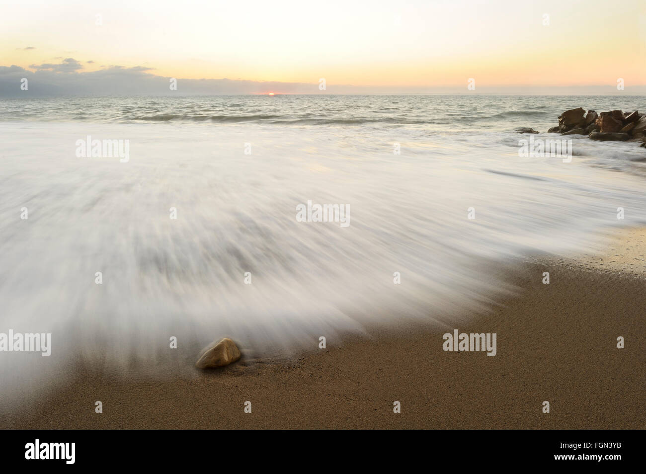 Ocean Sunset ist ein Ozean Rock an einem Sandstrand mit einer sanften Welle Rollen und einen weichen Pastell Sonnenuntergang am Horizont. Stockfoto