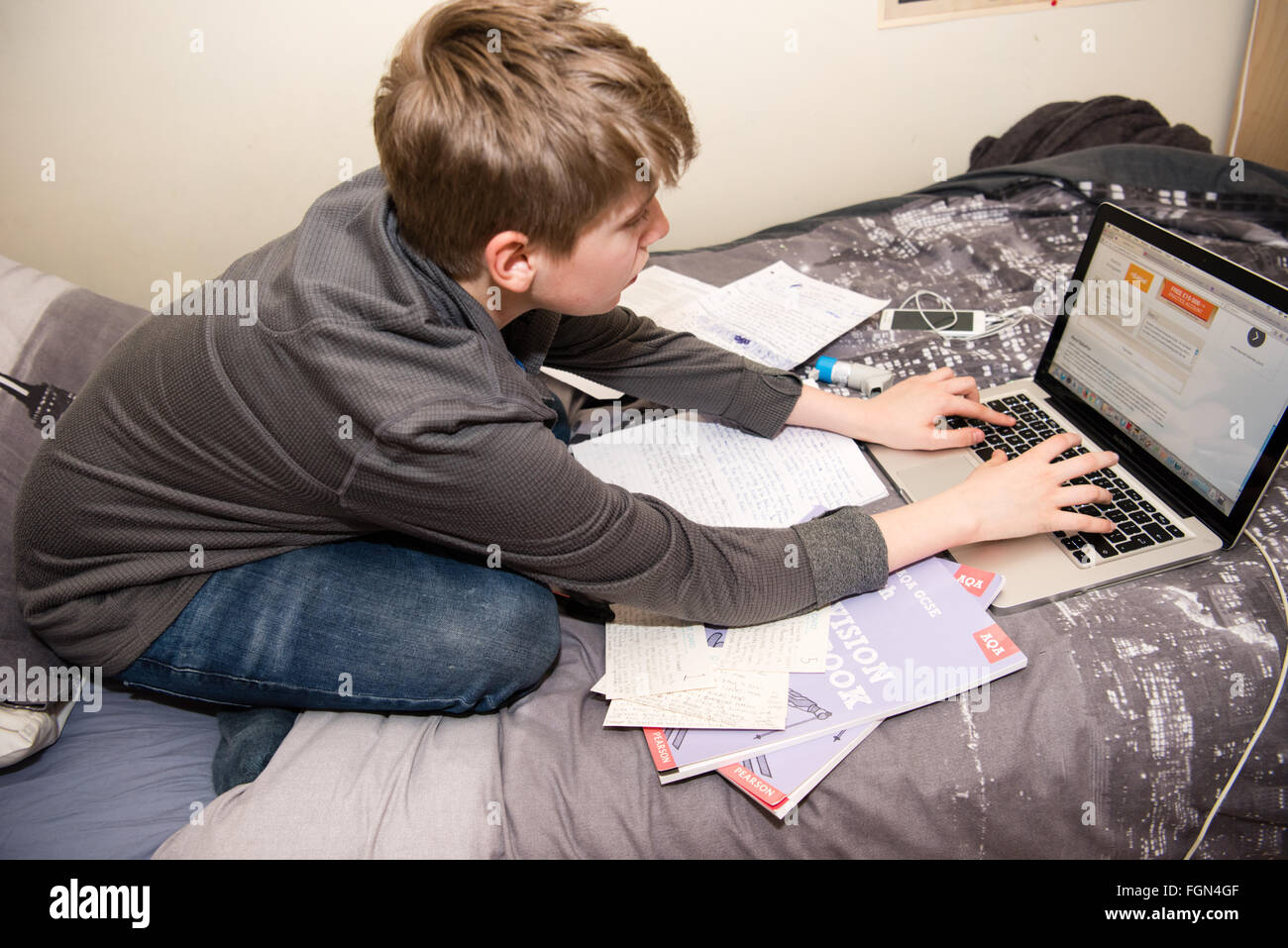 Weiße Teenager sitzen auf seinem Bett mit einem Laptop und viele Schulbücher, wie er für GCSE Prüfungen überarbeitet und Hinweis für seine Hausaufgaben macht. Stockfoto