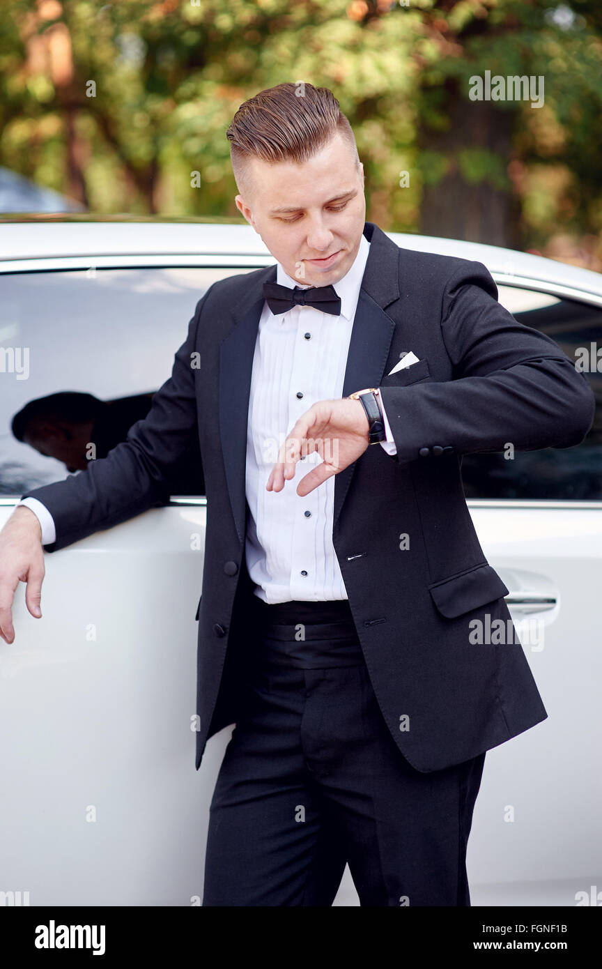 elegante stilvolle Bräutigam in der Nähe von Auto warten seine Brite und sieht auf Uhr Stockfoto