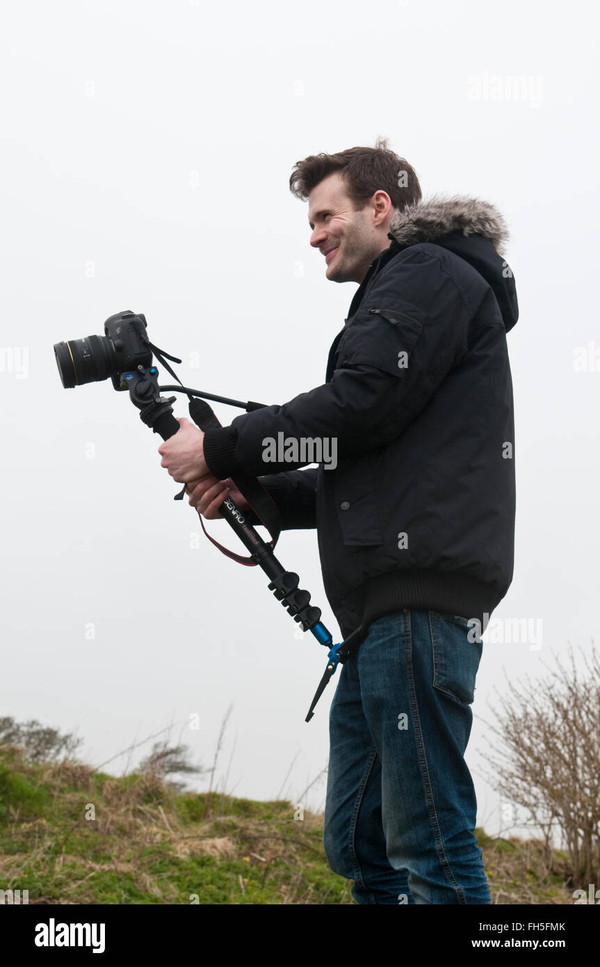 Junge Mann trägt einen schwarzen Parka Stil Mantel mit einer Canon EOS 5D DSLR mit einem Einbeinstativ als Videokamera Stockfoto