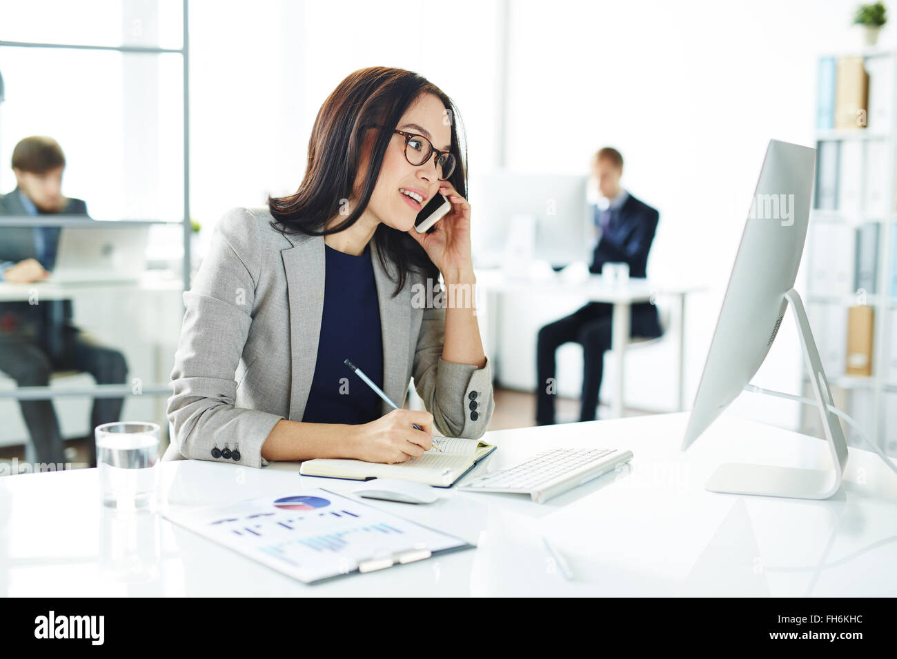 Beschäftigt Sekretär am Handy sprechen und Notizen im Notizbuch beim Monitor betrachten Stockfoto