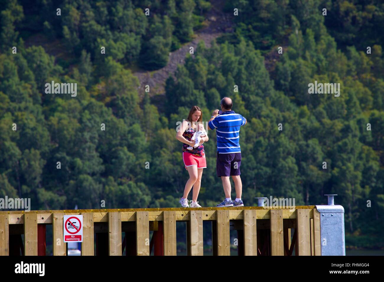 Lake District National Park. Mann nehmen Foto von seiner Frau und Baby mit Ihrem Smartphone auf einem Steg in Lake Ullswater. Stockfoto