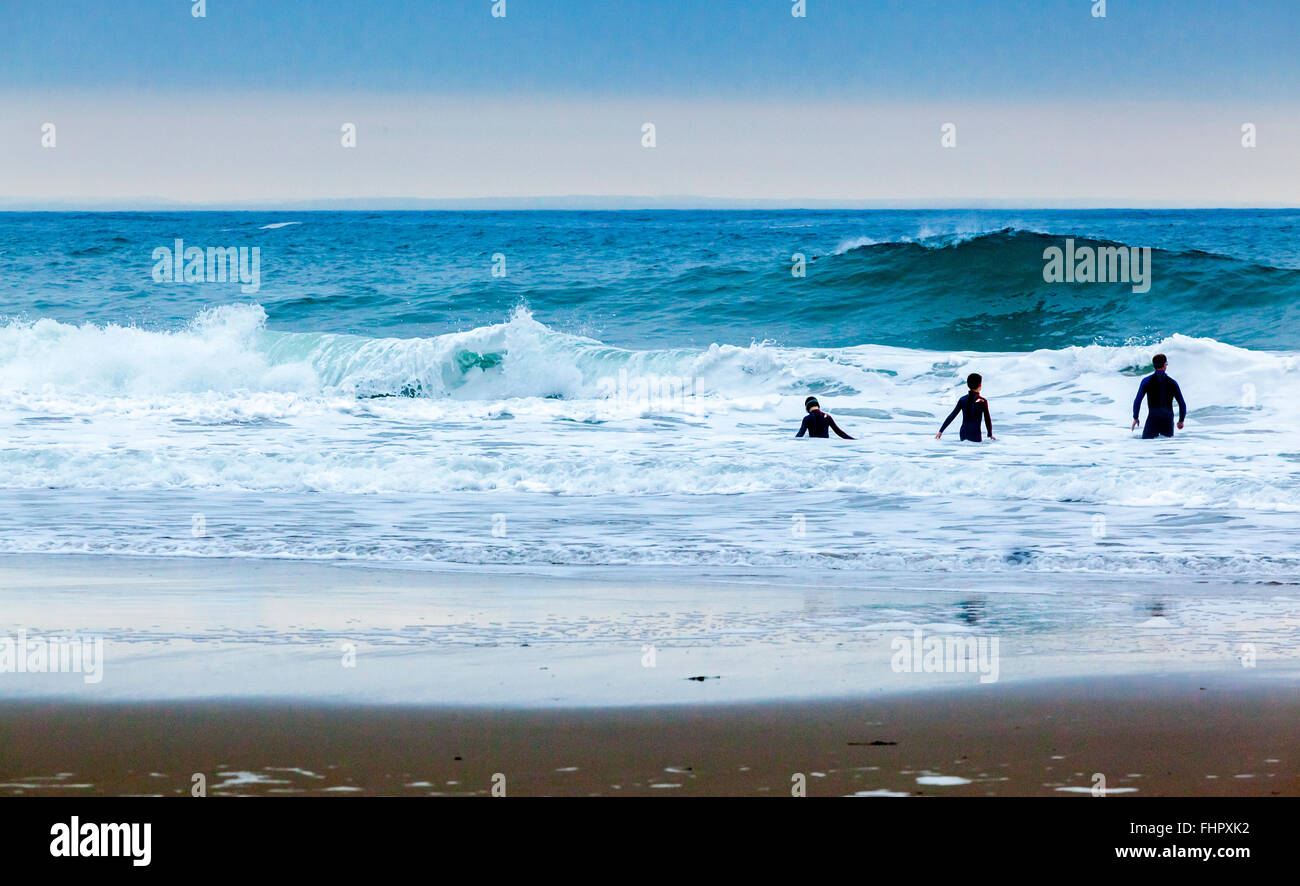 Silhouettenhafte Figuren von drei Personen spielen im Wasser an einem Strand mit Wellen, die hinter Stockfoto
