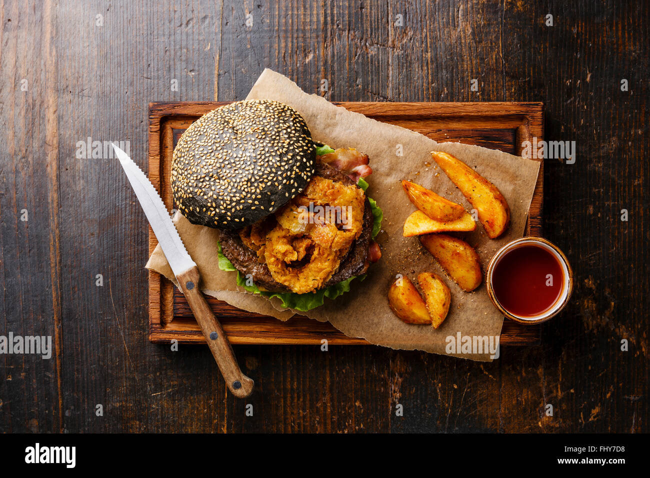 Schwarzen Burger mit Sesam Samen Brötchen Fleisch Speck Zwiebeln Pommes frites, Ringe und Kartoffelspalten auf dunklem Holz Stockfoto