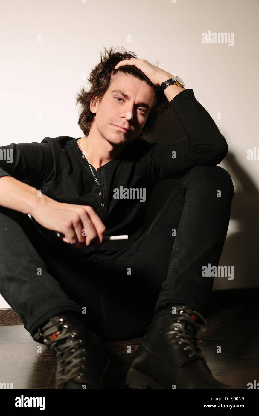 Junger Mann sitzt auf einer Etage einen Rücken zur Wand. Eine Hand auf den Kopf und ein anderes hält eine Zigarette. Stockfoto