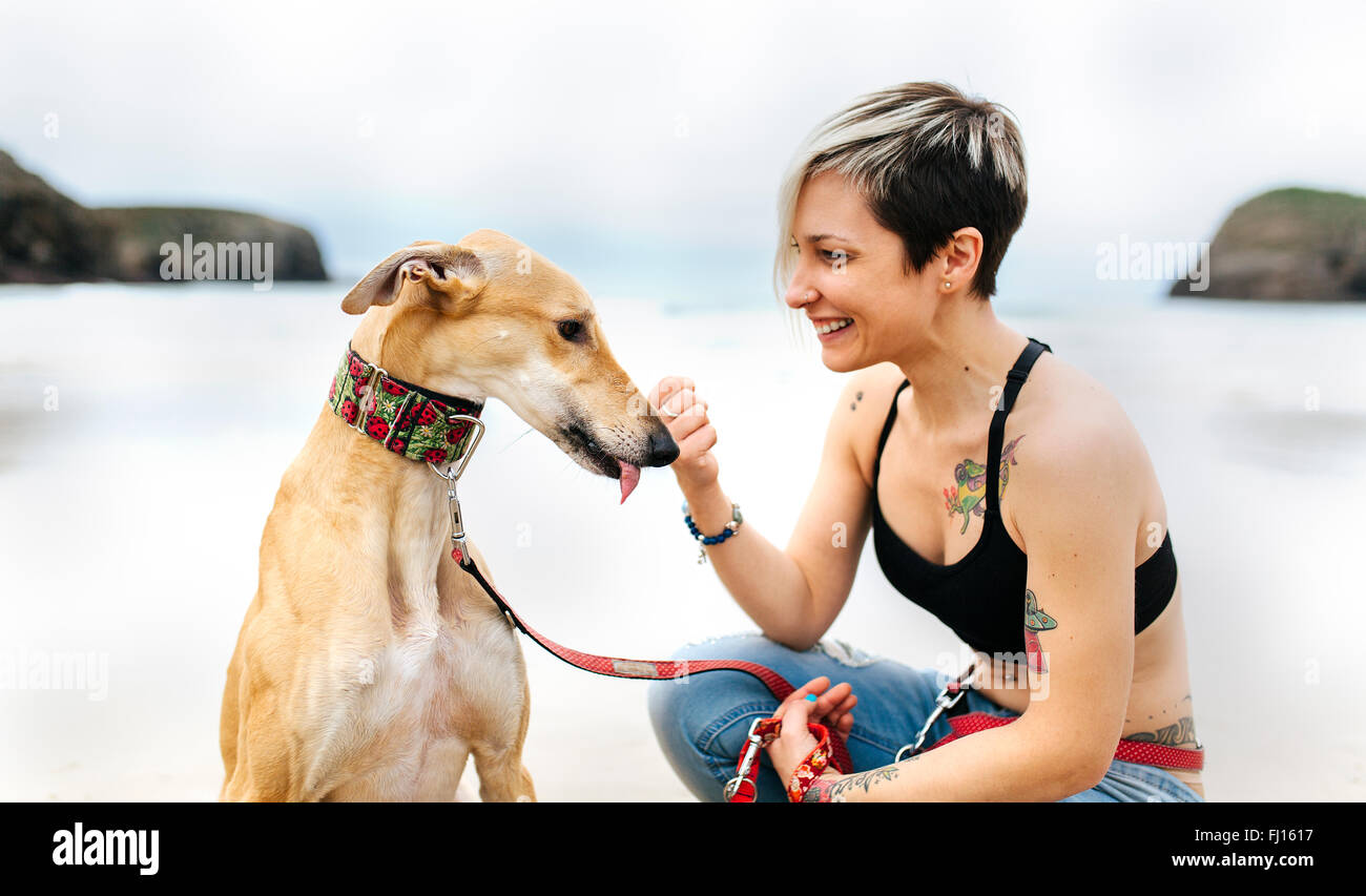 Spanien, Llanes, lächelnde junge Frau mit ihrem Windhund am Strand Stockfoto