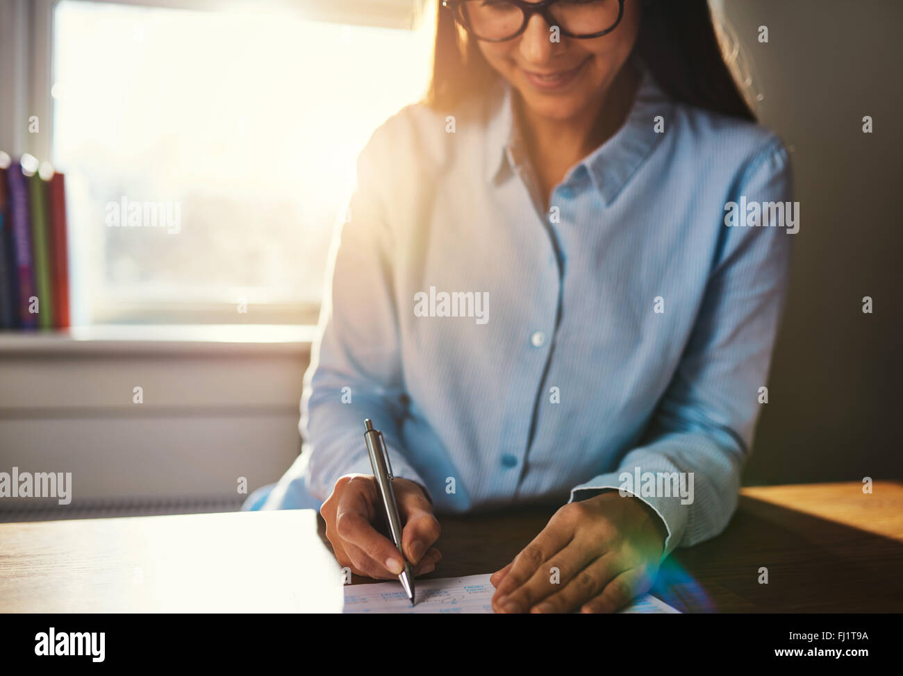 Selektiver Fokus auf die Hände von lächelnden Frau in Blau Bluse schreiben am Schreibtisch auf Notepad im Home Office mit Sonnenlicht über Ihr sollte Stockfoto