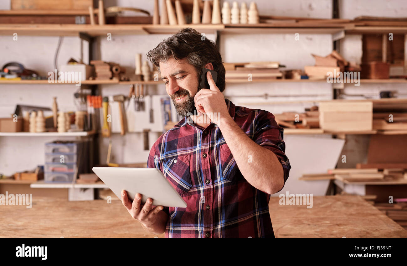 Mann mit Bart in Holzarbeiten Studio mithilfe von Handy und tablet Stockfoto