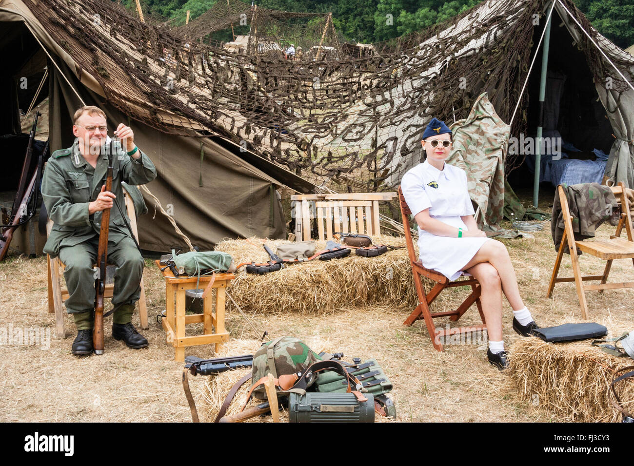 Zweiten Weltkrieg Re-enactment. Deutsche Camp mit Soldat sein Gewehr sitzend, Reinigung. Auf Stuhl eine Frau in weißen Uniform, Krankenschwester, Lächeln, Blickkontakt. Stockfoto