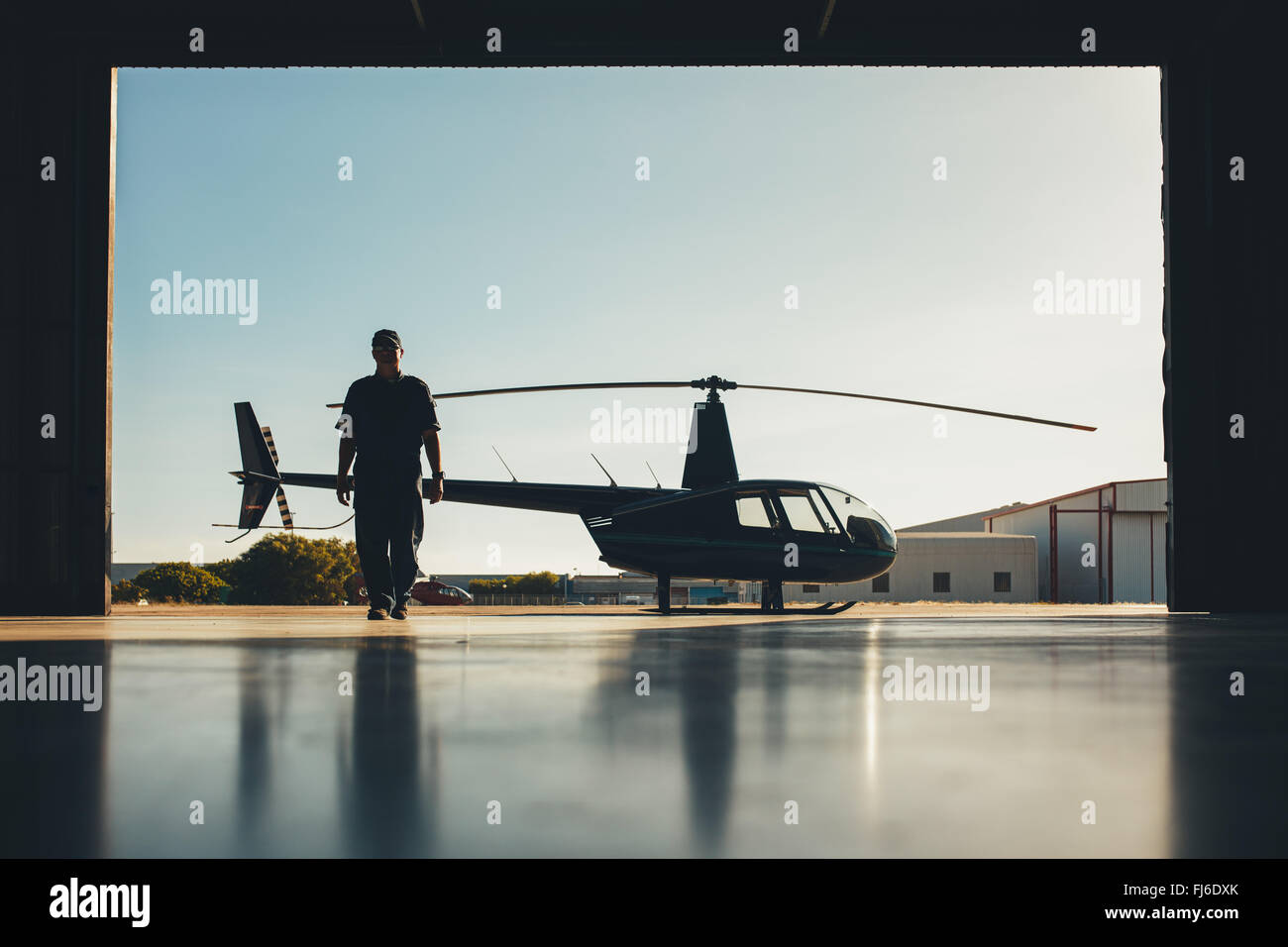 Silhouette der Hubschrauber mit einem Piloten im Flugzeughangar. Pilot zu Fuß entfernt von Hubschrauber vor der Halle geparkt. Stockfoto