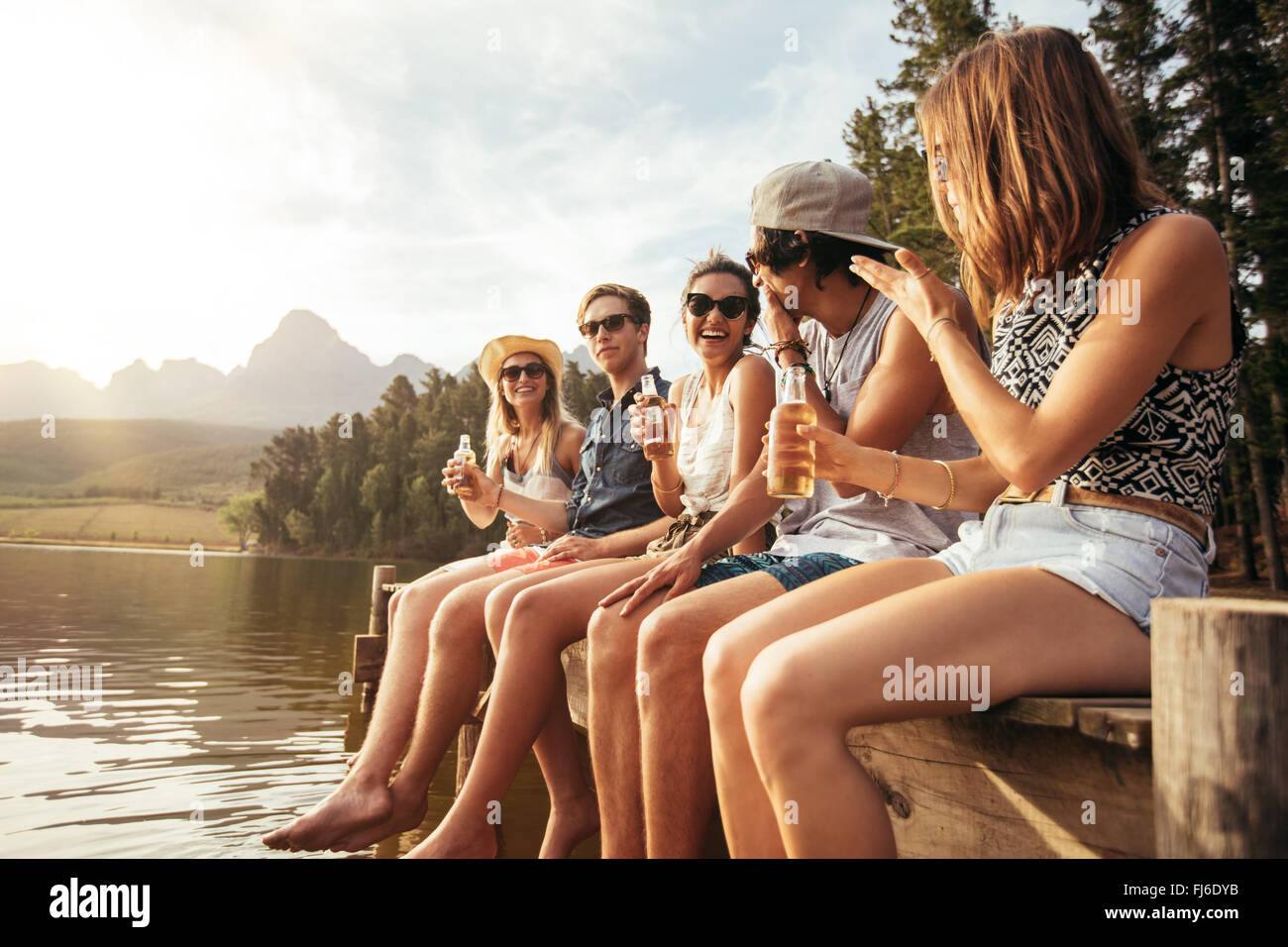 Porträt der glückliche junge Freunde sitzen auf Pier am See trinken Bier. Junge Männer und Frauen, die einen Tag am See zu genießen. Stockfoto