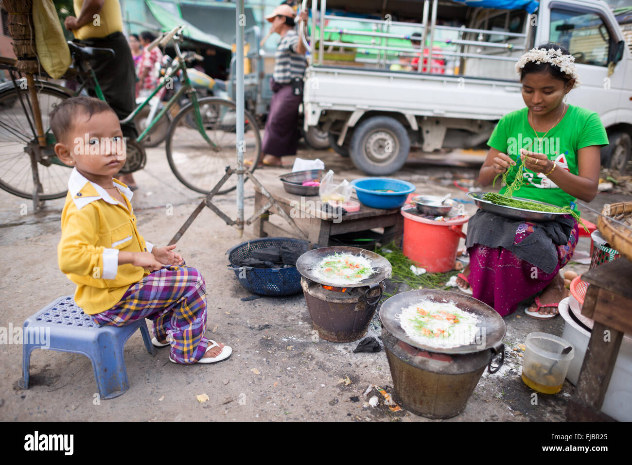 MANDALAY, Myanmar — Ein kleines Kind hilft ihrer Mutter, auf dem Fisch- und Blumenmarkt in Mandalay, Myanmar (Birma) Mahlzeiten zu kochen, die auf der Straße verkauft werden. Stockfoto