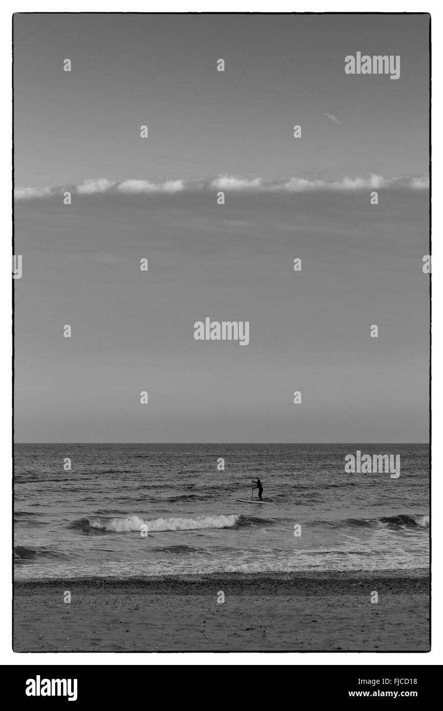 Goldene Abendlicht einer Silhouette eines Surfers auf seinem Brett, das Meer am Strand mit Sand und keine Wolken in Richtung der Stockfoto