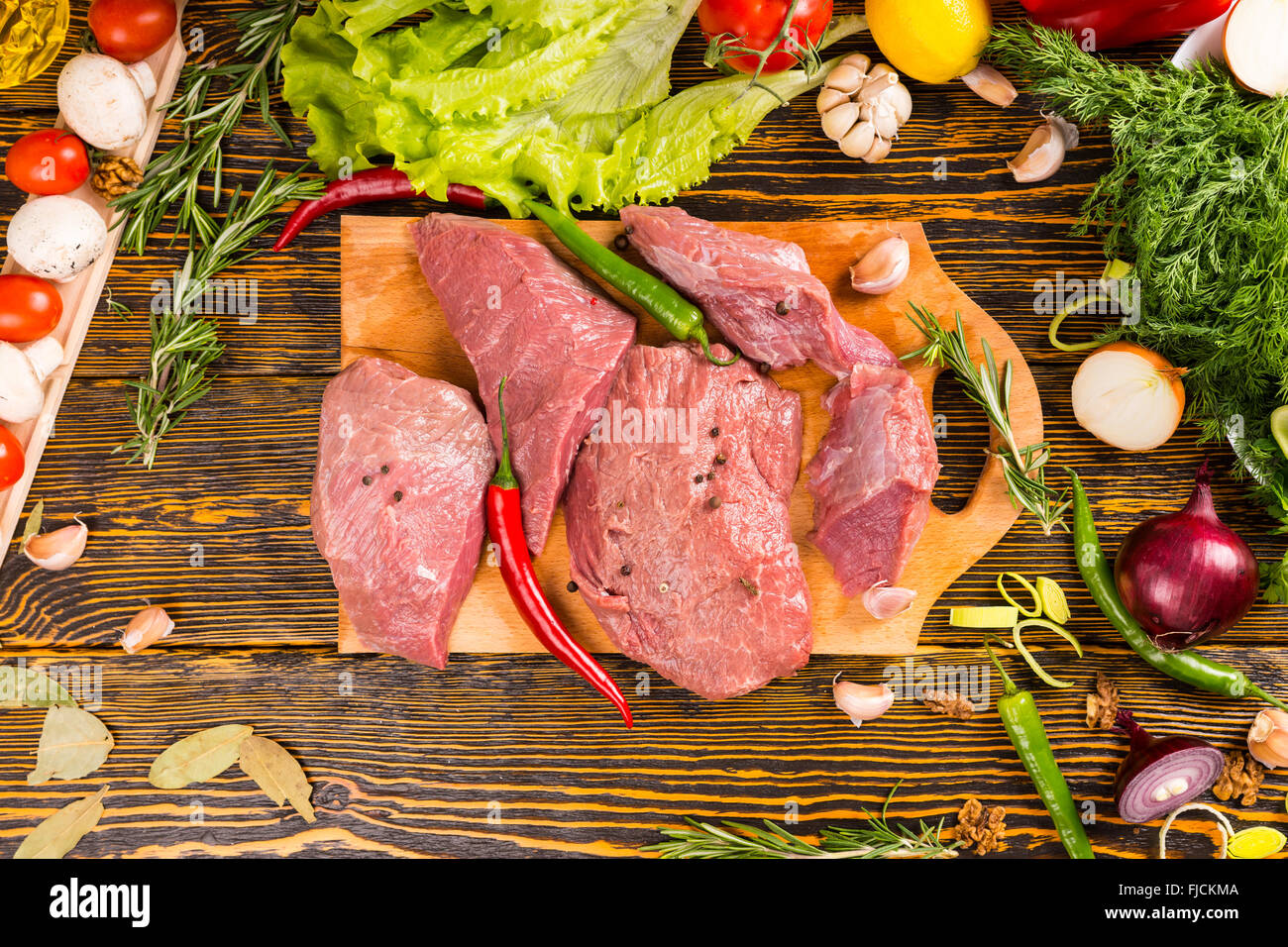 Über Blick über vier Schnitten von rohes, rotes Fleisch mit frischen Peperoni über Holztisch umgeben von Zwiebeln, Tomaten, Pilze und anderes Gemüse. Stockfoto