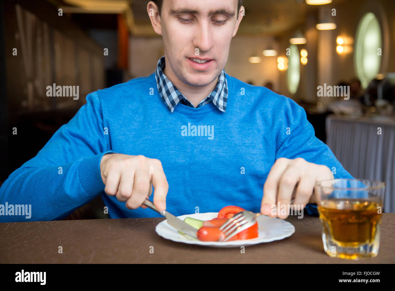 Porträt von lächelnden jungen Mann drinnen essen Gemüse mit Wurst mit Messer und Gabel Stockfoto