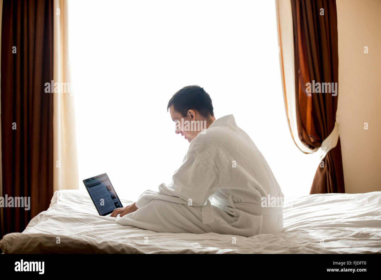 Porträt der attraktive junge Mann sitzt auf Bett mit weißen Bademantel, konzentrierte sich auf die Arbeit, männliches Model mit Laptop im hotel Stockfoto