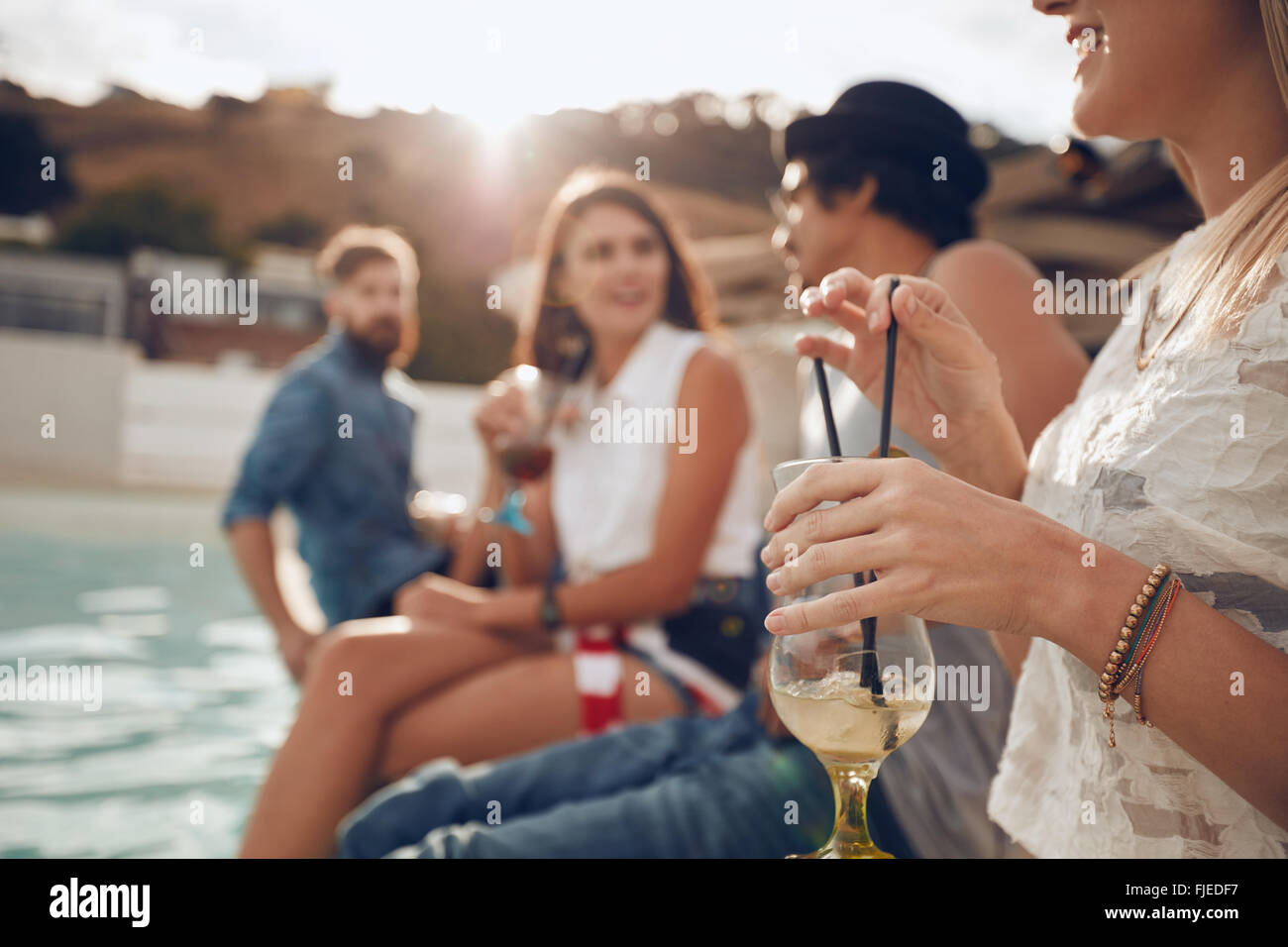 Frau hält ein Cocktailglas sitzend am Rand des Swimming Pool mit Freunden. Junge Menschen genießen eine Pool-Party-w Stockfoto