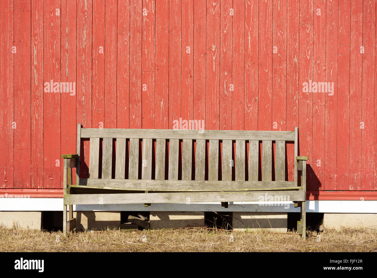 Eine alte unlackiert Bank gegen eine rote Holzwand. Sonne strahlt auf die leere outdoor-Möbel. Kopieren Sie Platz an Wand. Stockfoto