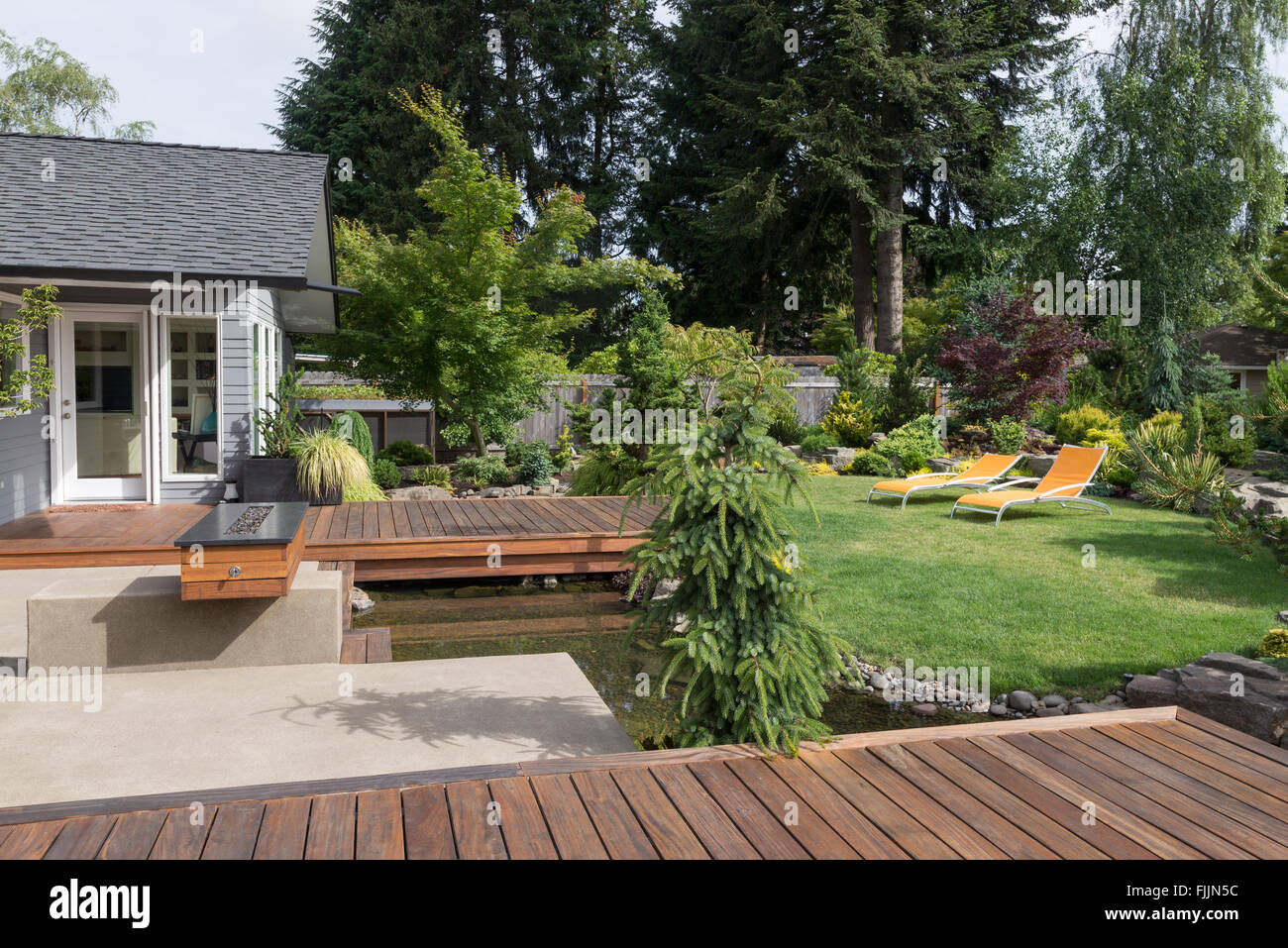 Hinterhof eines modernen pazifischen Nordwesten-Hauses mit einem Deck über einen Bach erinnernde Wasserspiel mit einem gepflegten Rasen. Stockfoto