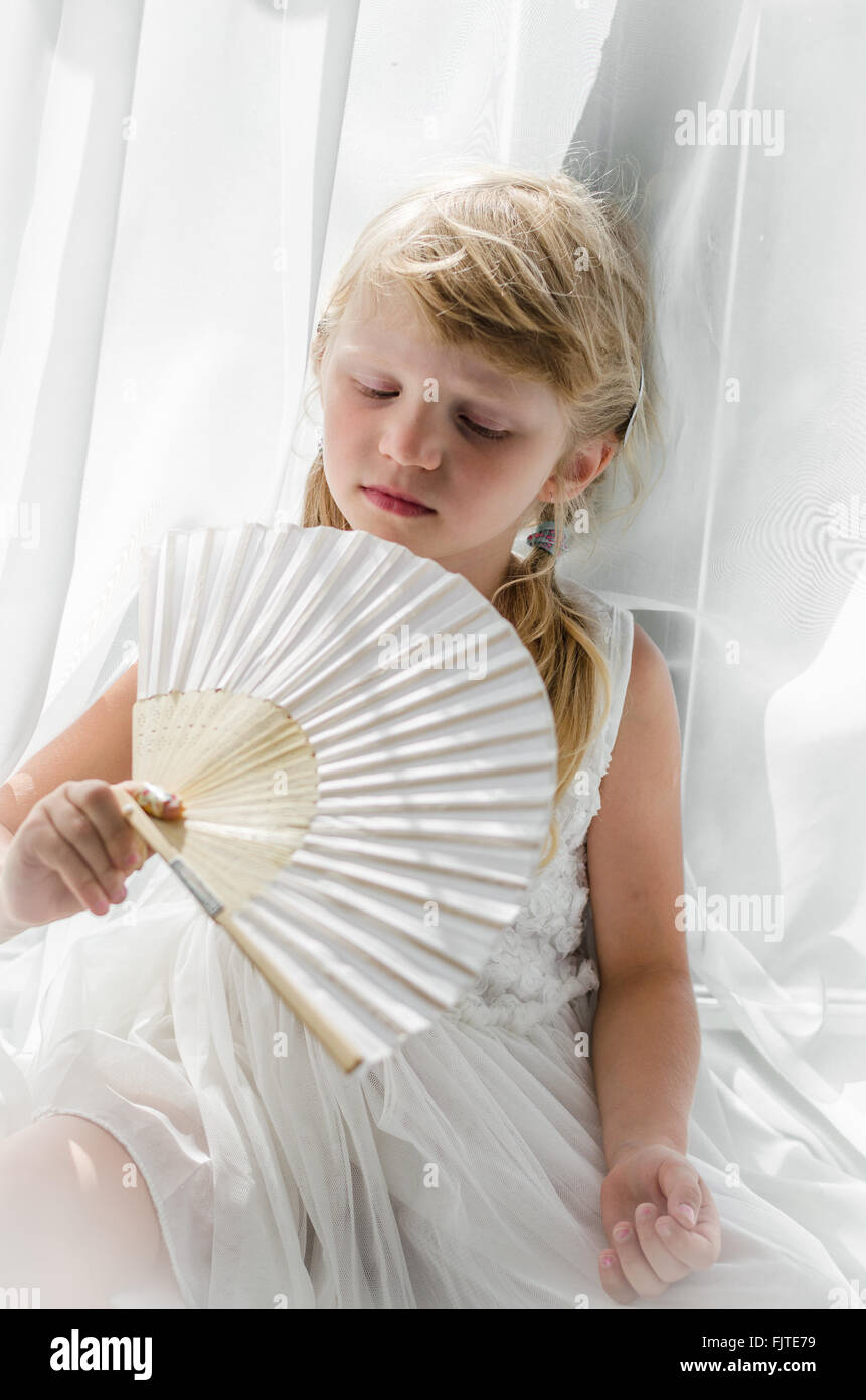 unschuldige niedliche kleine Mädchen sitzen durch den Vorhang und halten weiße Lüfter Stockfoto