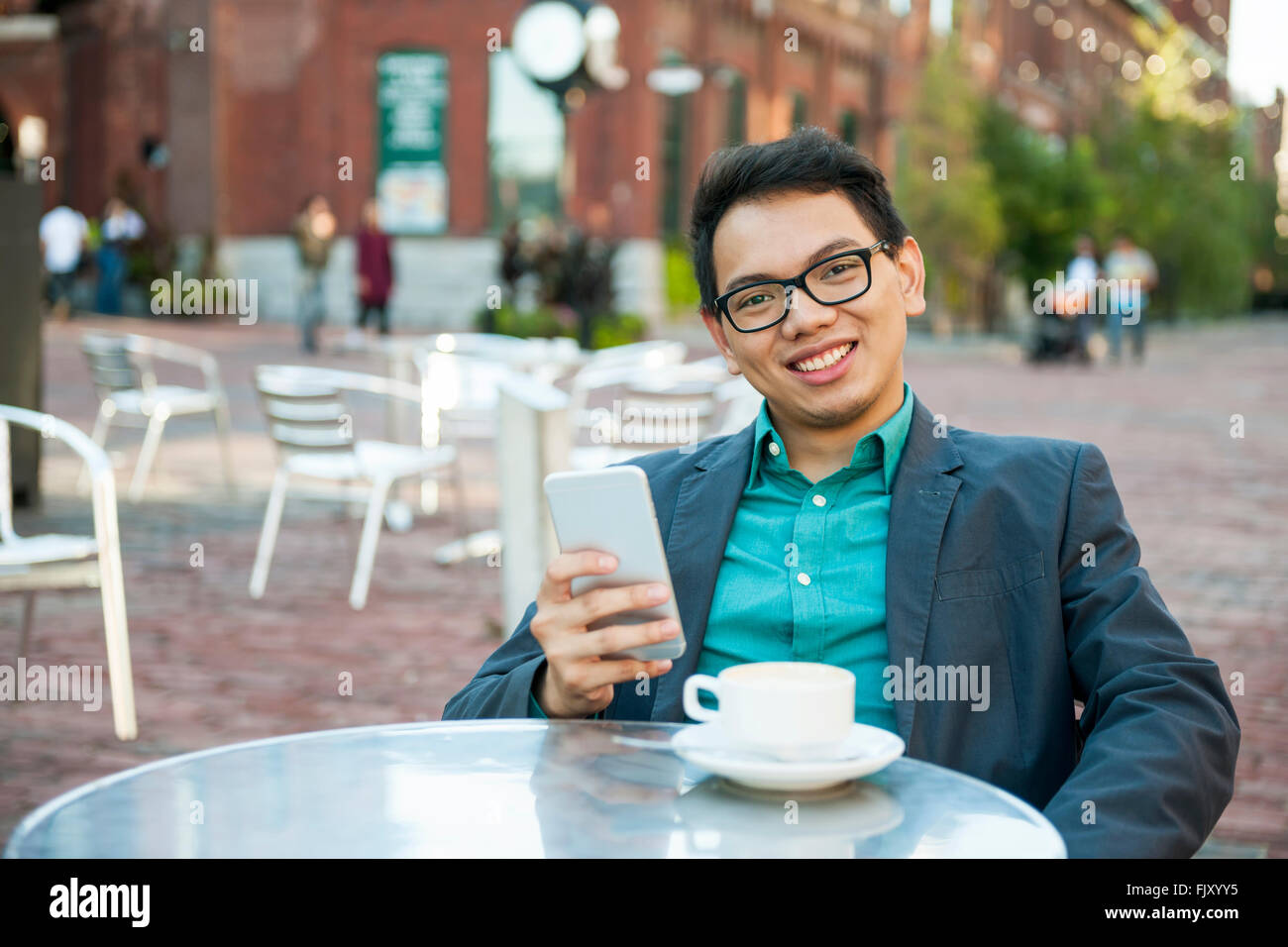 Junge asiatische Mann in casual Geschäftskleidung lächelnd in entspannten outdoor-Café mit Kaffee und Handy Stockfoto