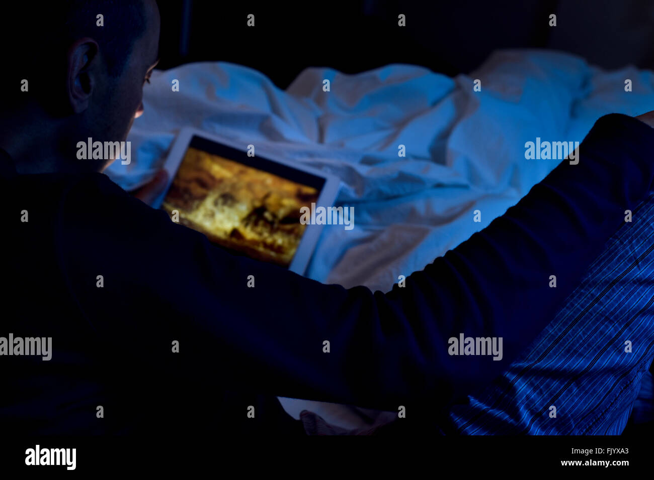 ein junger kaukasischer Mann auf dem Bett liegend Uhren einen Film oder eine Serie in streaming auf seinem Tablettcomputer in der Nacht Stockfoto