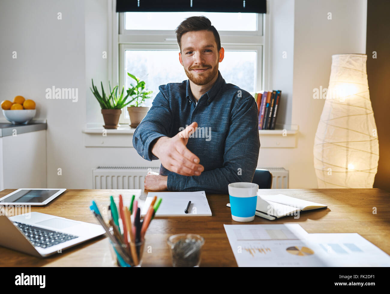 Lächelnd Geschäftsmann lehnt sich auf seinem Schreibtisch bietet seine Hand in Gruß, ein Geschäft in Partnerschaft oder Herzlichen Glückwunsch schließen Stockfoto