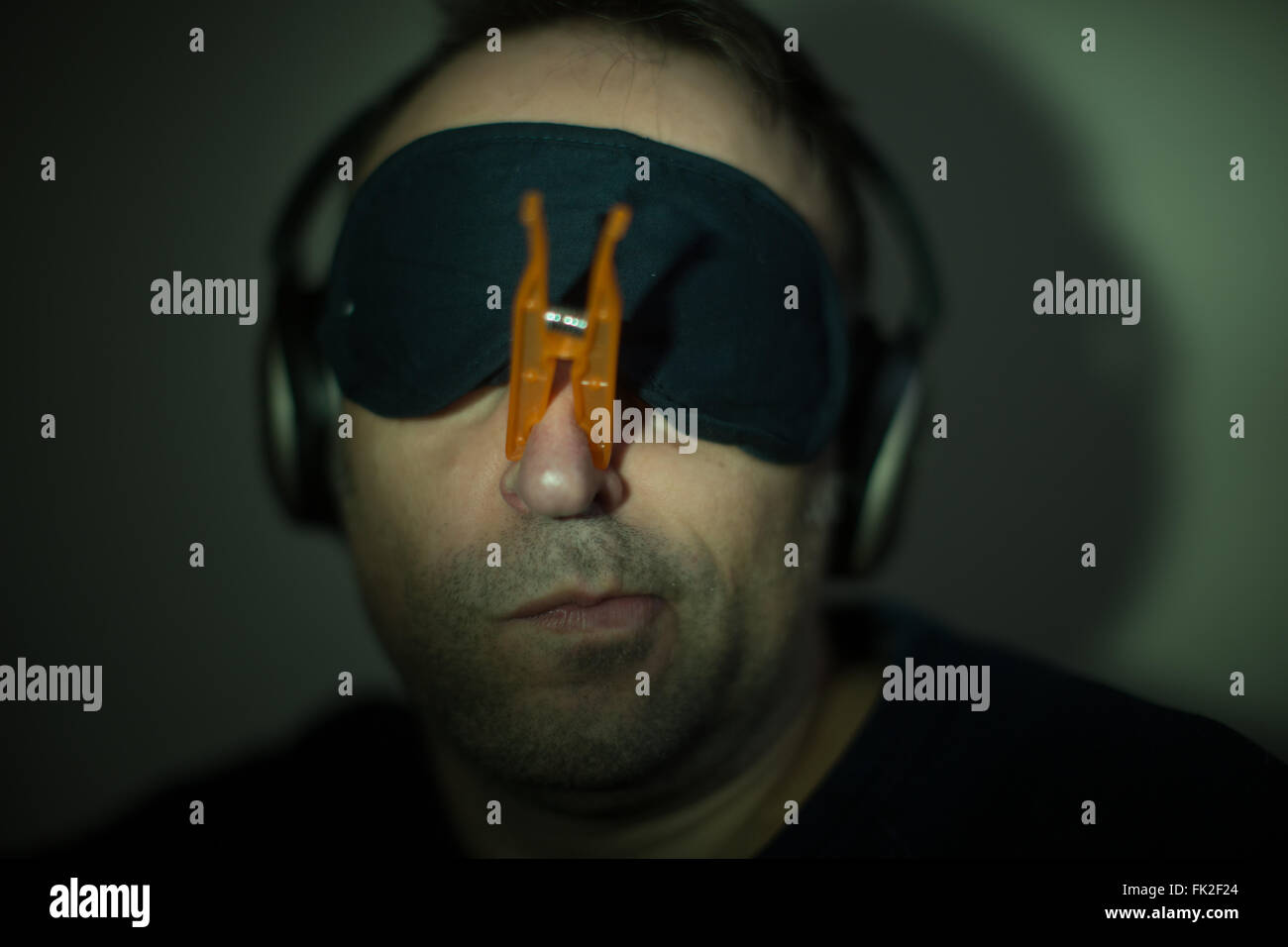 Ein Mann trägt eine Wäscheklammer auf der Nase, Kopfhörer und eine Augenmaske Stockfoto