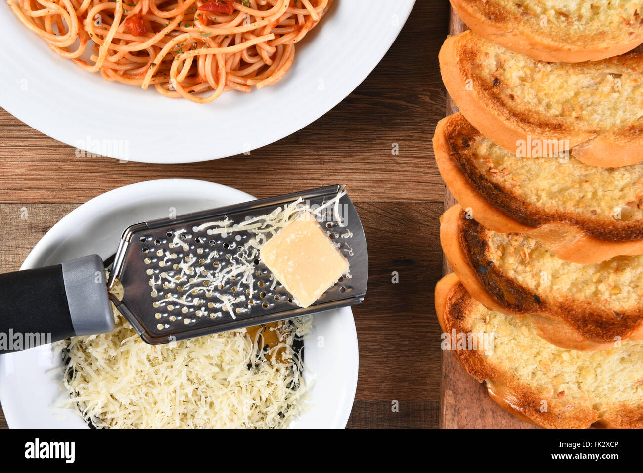 Draufsicht auf Knoblauchbrot auf ein Schneidebrett und einen Teller Spaghetti mit einer Käsereibe mit geriebenem Parmesan. Stockfoto