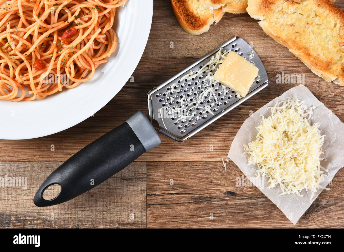 Erhöhte Ansicht einer Käse reiben mit Parmesan-Käse auf einem rustikalen Holz Küchentisch. Einen Teller mit Spaghetti und Knoblauch Brot ar Stockfoto